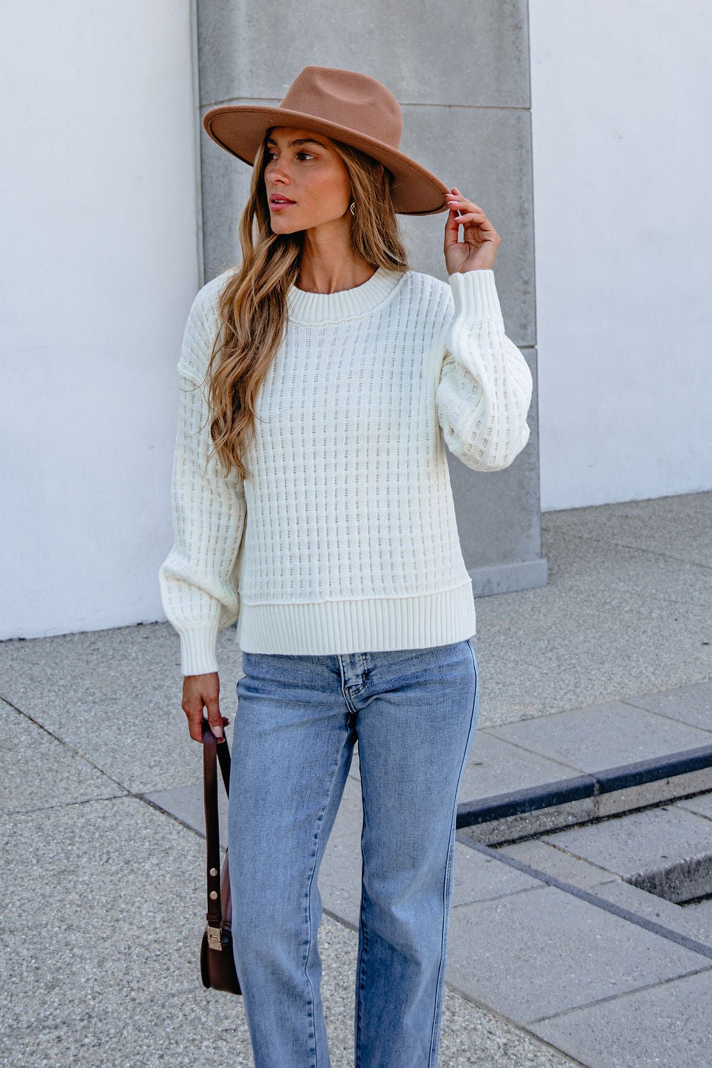 Woman in cream waffle knit sweater, brown hat, and light blue jeans stands outdoors on a concrete surface.