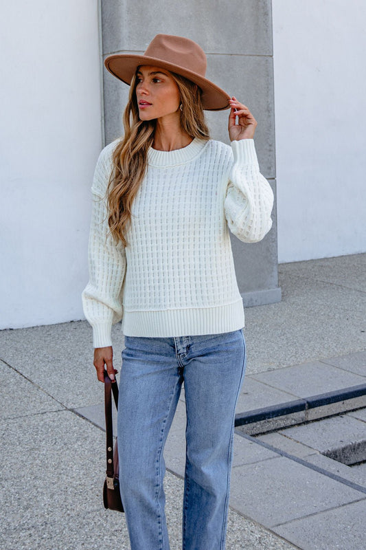 Woman in cream waffle knit sweater, brown hat, and light blue jeans stands outdoors on a concrete surface.