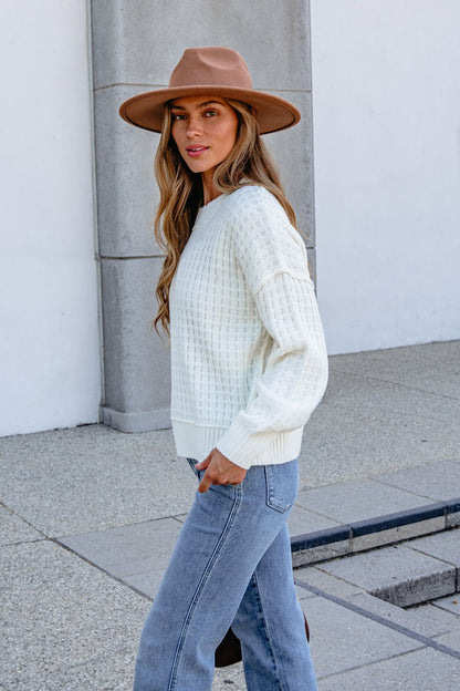 Woman in a Cream Waffle Knit Sweater, brown hat, and blue jeans walks outside on a paved sidewalk.