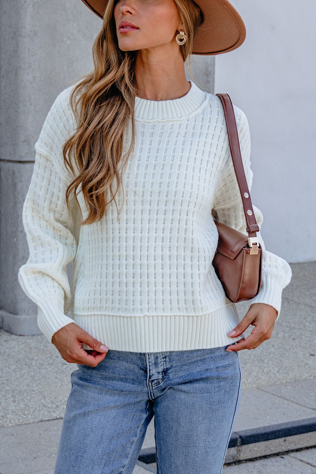 Woman in a Cream Waffle Knit Sweater, jeans, tan hat, and brown shoulder bag stands outdoors.