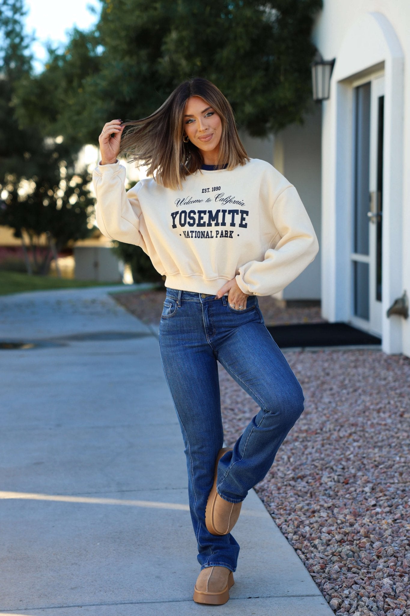 A woman smiles outside in a Cream Yosemite Graphic Crewneck Pullover, jeans, and boots, standing on the sidewalk holding her hair.