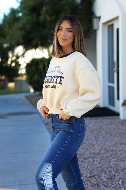 Woman with straight brown hair wears a Cream Yosemite Graphic Crewneck Pullover and blue jeans outdoors on a sunny day.
