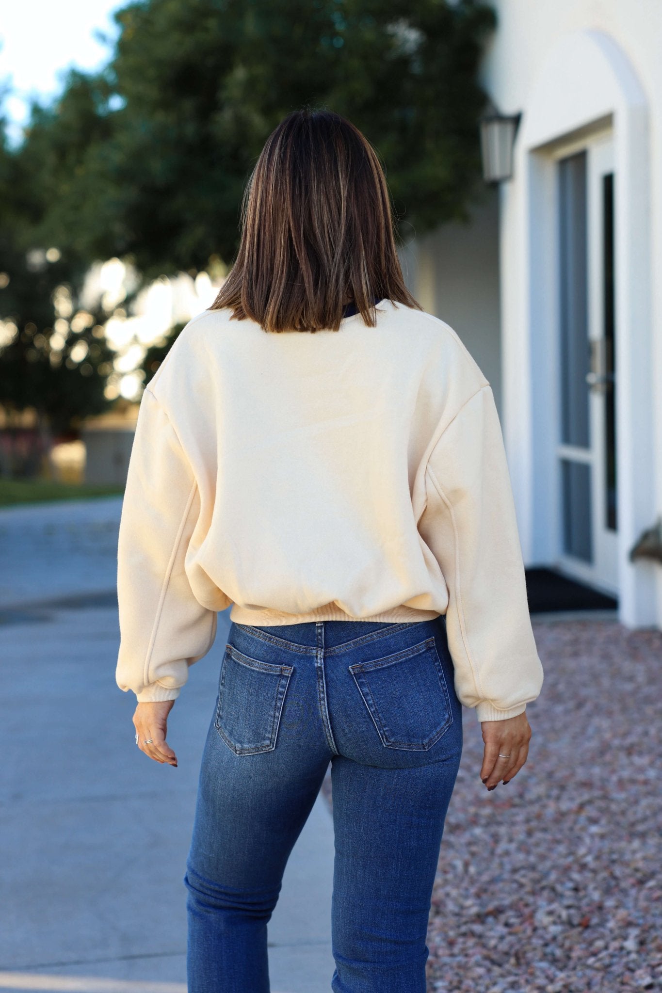 A woman in a Cream Yosemite Graphic Crewneck Pullover and blue jeans stands outdoors, facing away with brown hair loose.