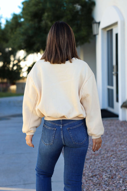A woman in a Cream Yosemite Graphic Crewneck Pullover and blue jeans stands outdoors, facing away with brown hair loose.
