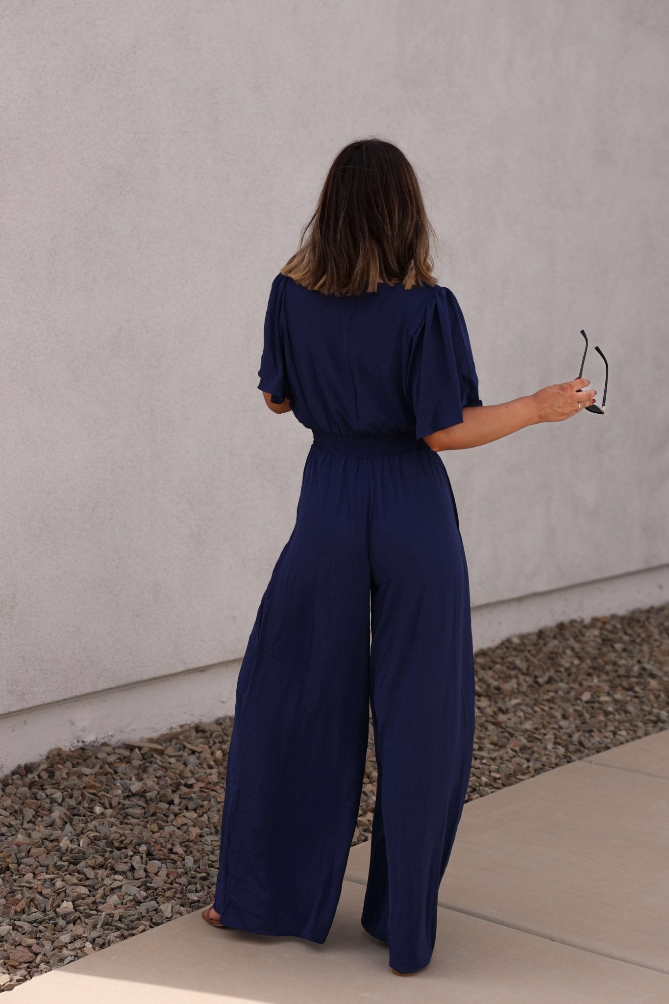 A woman stands barefoot outdoors, facing away, wearing the Crisp Horizon Navy Smocked Jumpsuit and holding sunglasses.