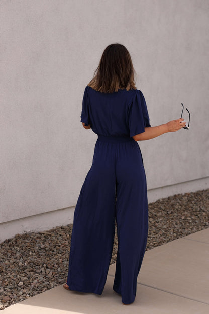 A woman stands barefoot outdoors, facing away, wearing the Crisp Horizon Navy Smocked Jumpsuit and holding sunglasses.