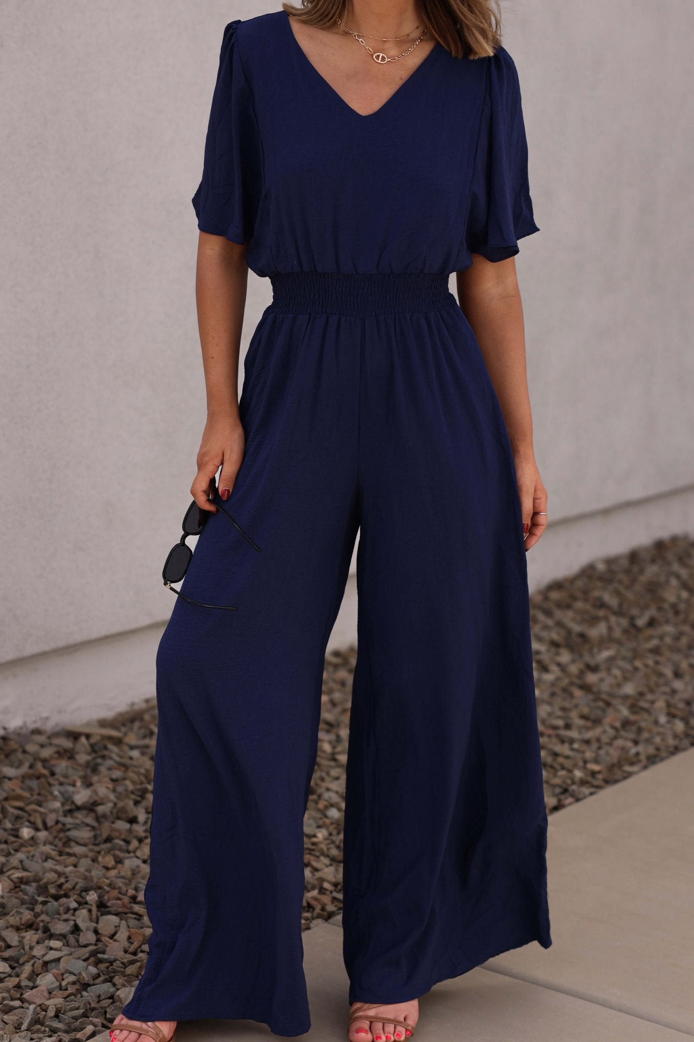 Woman in the Crisp Horizon Navy Smocked Jumpsuit stands on a sidewalk with sunglasses near a gravel area. FINAL SALE.