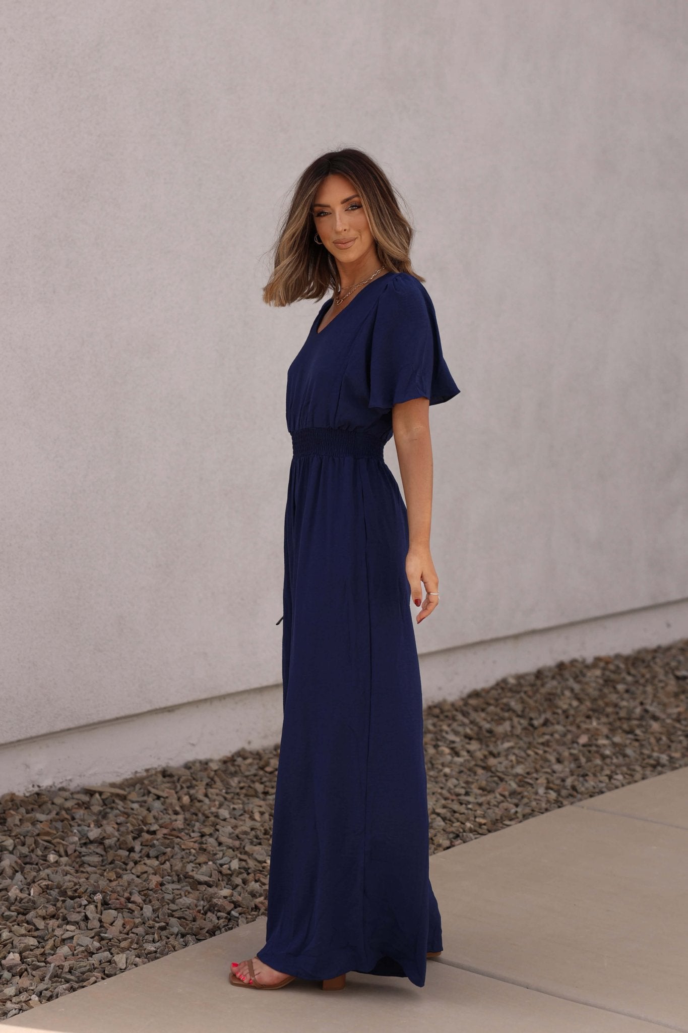 Woman wears the Crisp Horizon Navy Smocked Jumpsuit, standing sideways outdoors with a neutral wall and rocks behind her.