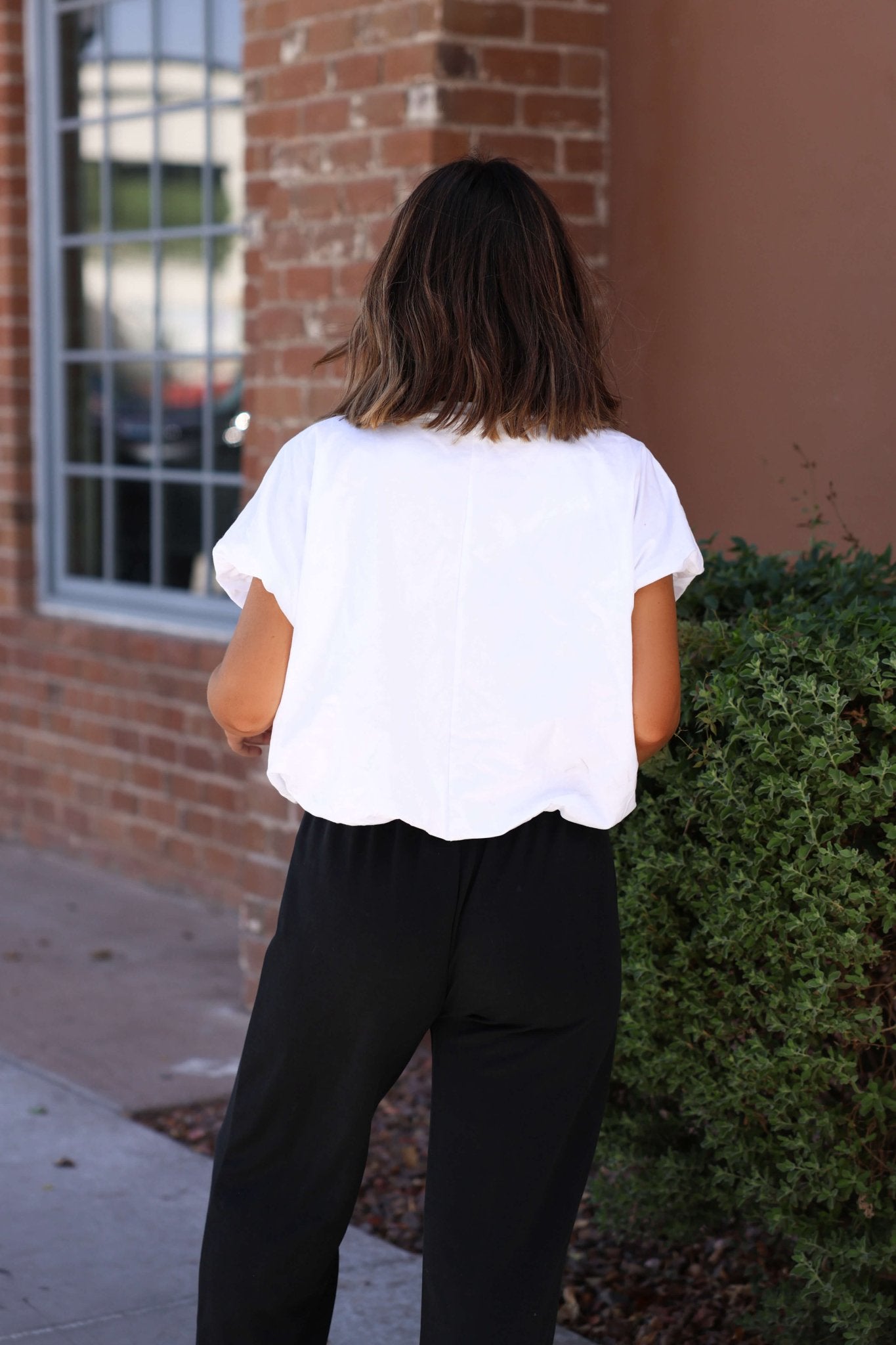 A woman with shoulder-length brown hair in the Crisp Morning Bubble Hem Top - White and black pants stands by a brick wall and bush.