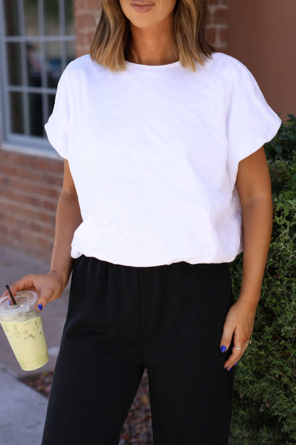 Woman in a Crisp Morning Bubble Hem Top - White and black pants holds an iced drink by greenery and a brick wall.