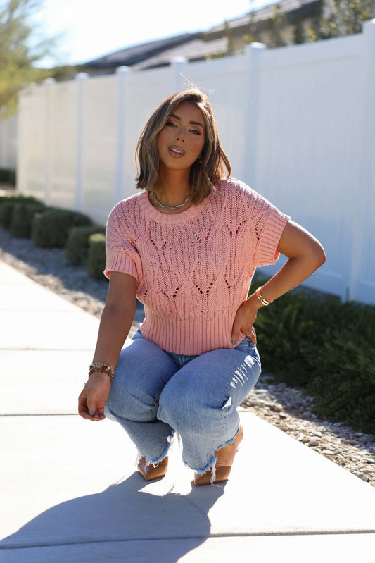 Woman in a Crochet Detail Short Sleeve Sweater - Blush, squats on a sunny sidewalk near a white fence and green shrubs.