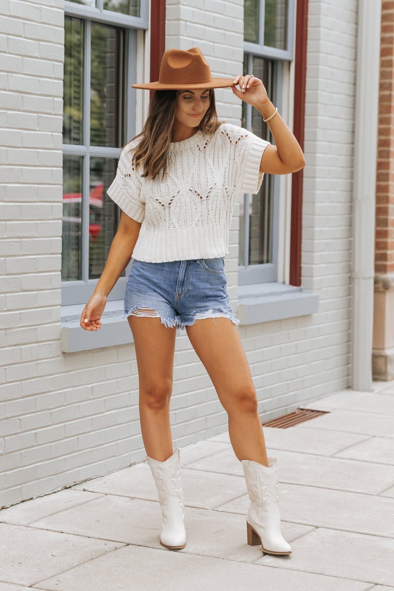 A woman in an ivory crochet detail short sleeve sweater, denim shorts, and cowboy boots stands by a brick building.