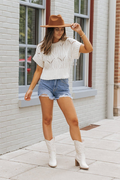 A woman in an ivory crochet detail short sleeve sweater, denim shorts, and cowboy boots stands by a brick building.