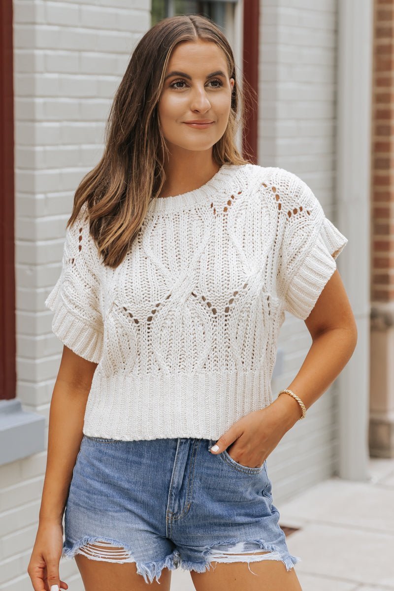 Woman wears an ivory Crochet Detail Short Sleeve Sweater with distressed denim shorts, standing by a brick wall.