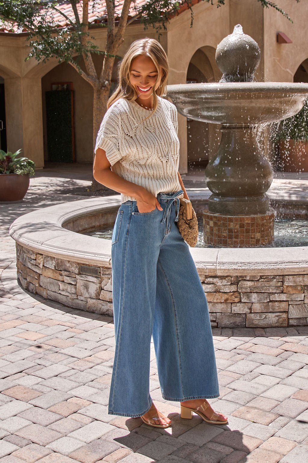 Woman smiling outdoors by a fountain, wearing the Crochet Detail Short Sleeve Sweater in Ivory and holding a woven clutch.