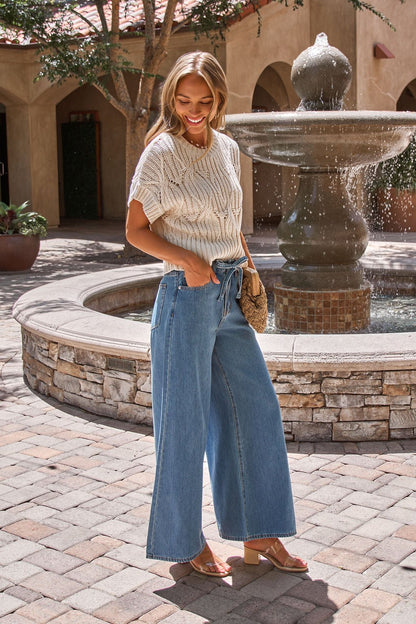 Woman smiling outdoors by a fountain, wearing the Crochet Detail Short Sleeve Sweater in Ivory and holding a woven clutch.