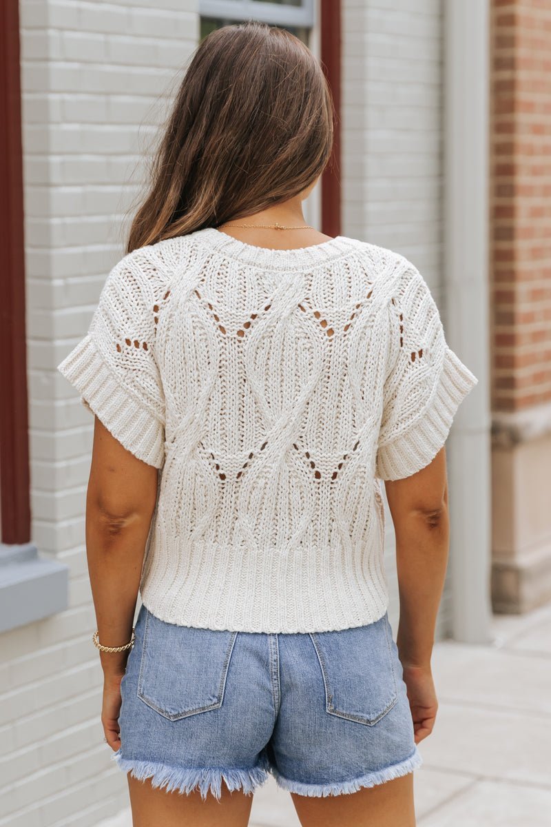 Woman with long brown hair wears the Crochet Detail Short Sleeve Sweater - Ivory and denim shorts, outdoors with her back to the camera.