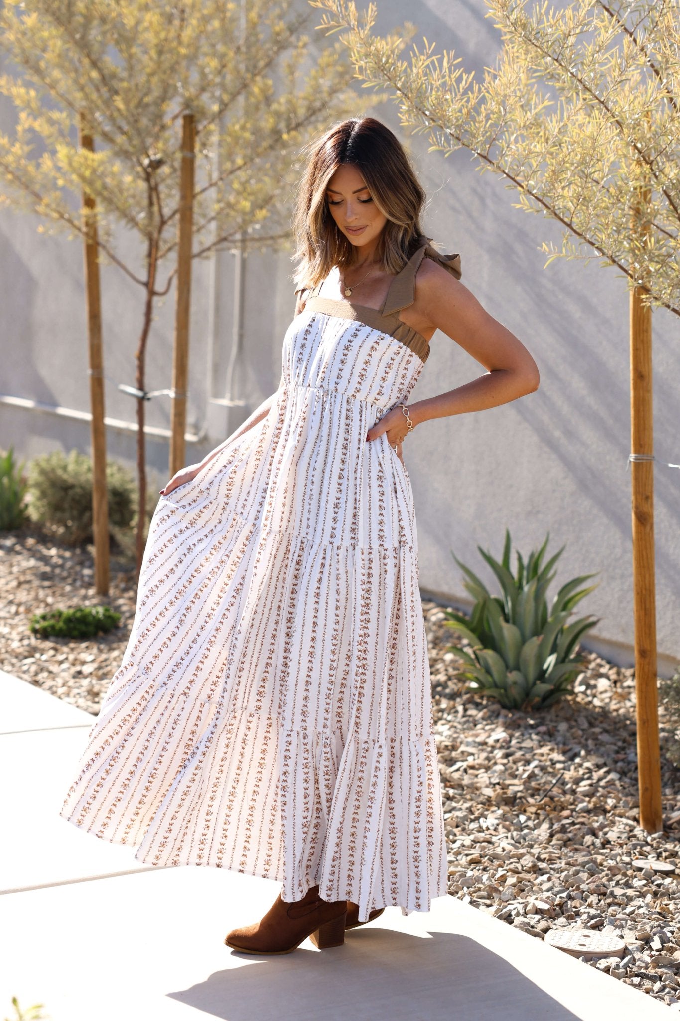 A woman in the Dahlia Bloom Ivory Floral Tiered Maxi Dress stands outdoors in brown boots, surrounded by greenery.