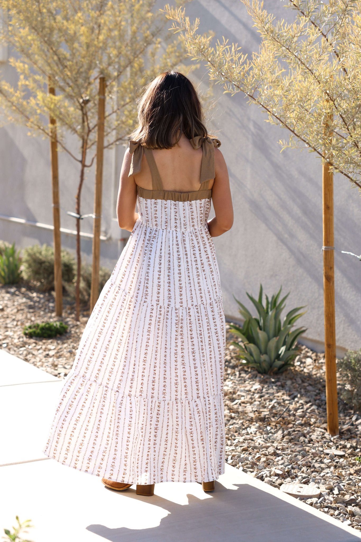 Woman wearing the Dahlia Bloom Ivory Floral Tiered Maxi Dress walks a sunlit path lined with trees and desert plants, back to camera.