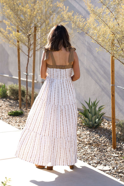 Woman wearing the Dahlia Bloom Ivory Floral Tiered Maxi Dress walks a sunlit path lined with trees and desert plants, back to camera.