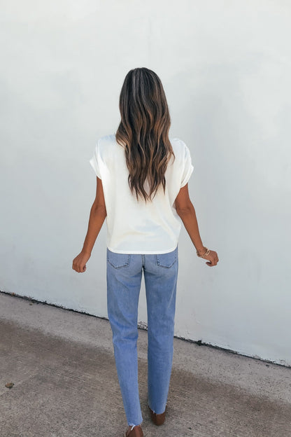 Woman with long wavy hair in the Daily Ruched Top - Ivory and blue jeans stands facing a white wall, arms slightly bent.