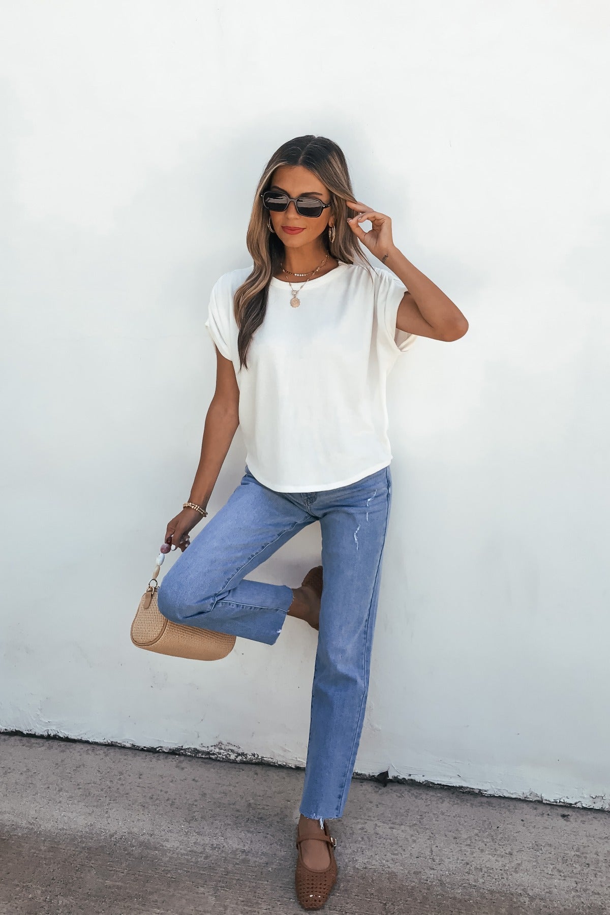 Woman in sunglasses, relaxed fit jeans, Daily Ruched Top - Ivory, and brown shoes poses with a beige bag against a white wall.