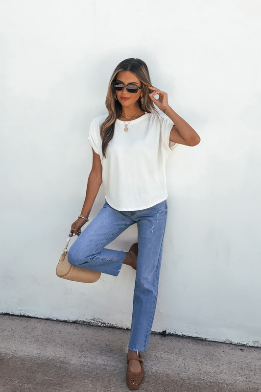 Woman in sunglasses, relaxed fit jeans, Daily Ruched Top - Ivory, and brown shoes poses with a beige bag against a white wall.