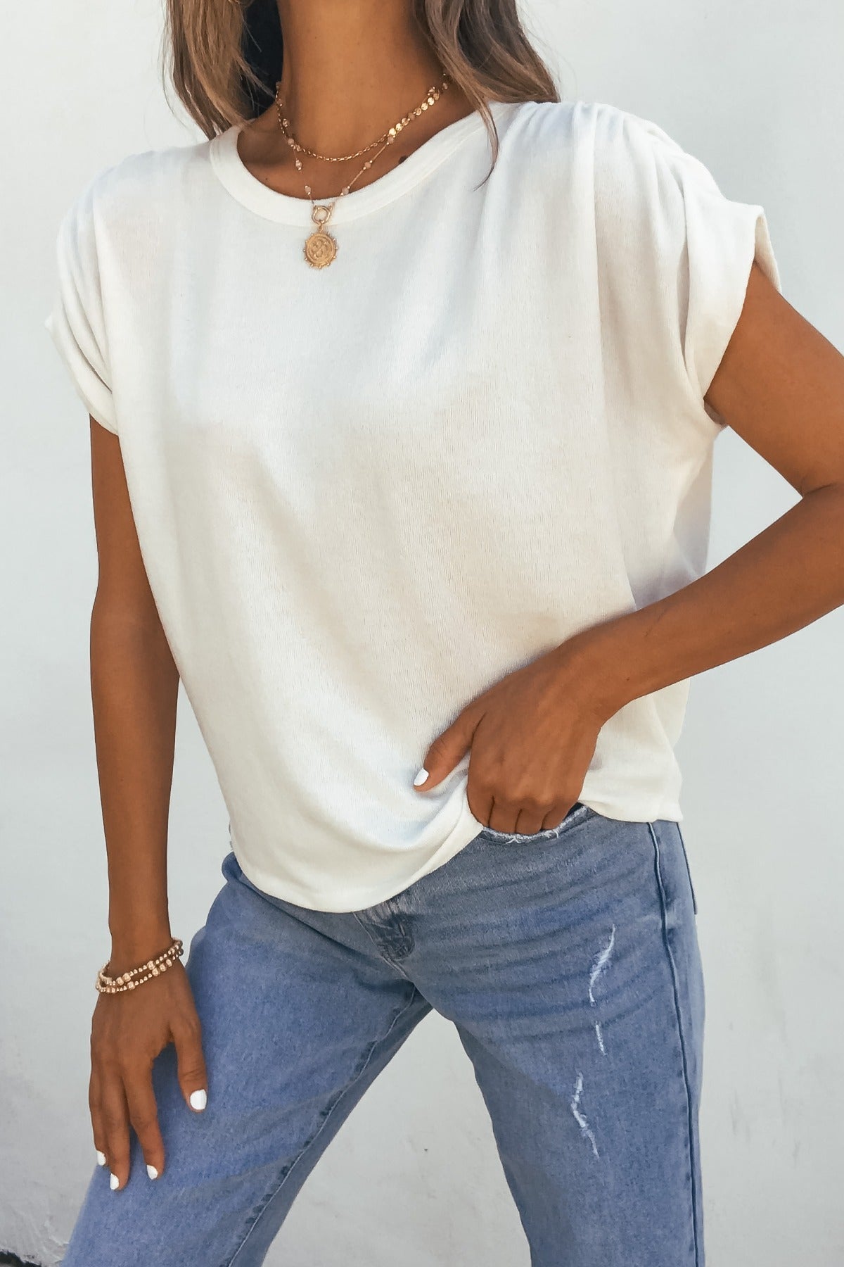 Woman wearing the Daily Ruched Top - Ivory with light blue jeans, necklaces, and bracelets, standing against a white wall.
