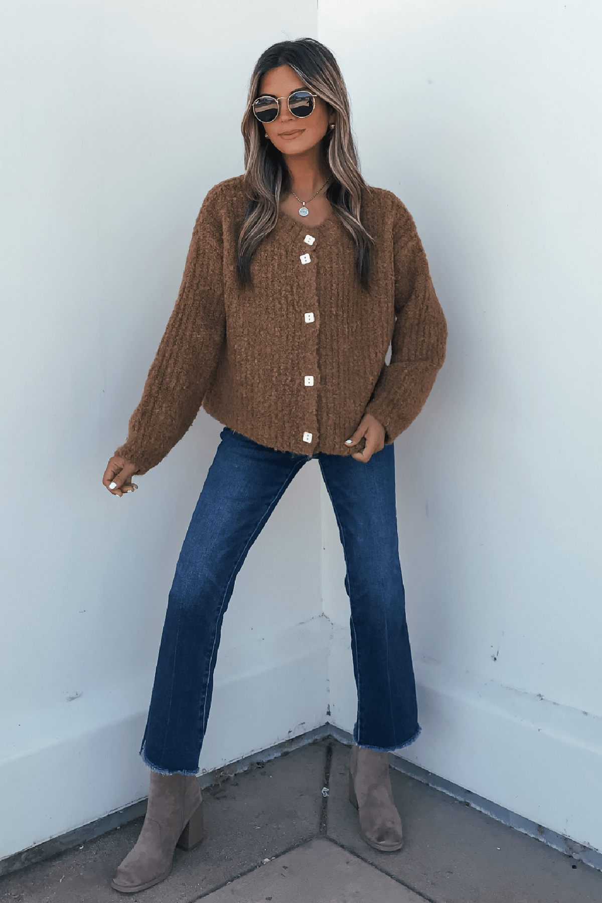 Woman in sunglasses, Dark Amber Plush Button Down Cardigan, blue jeans, and taupe ankle boots stands by a white wall.