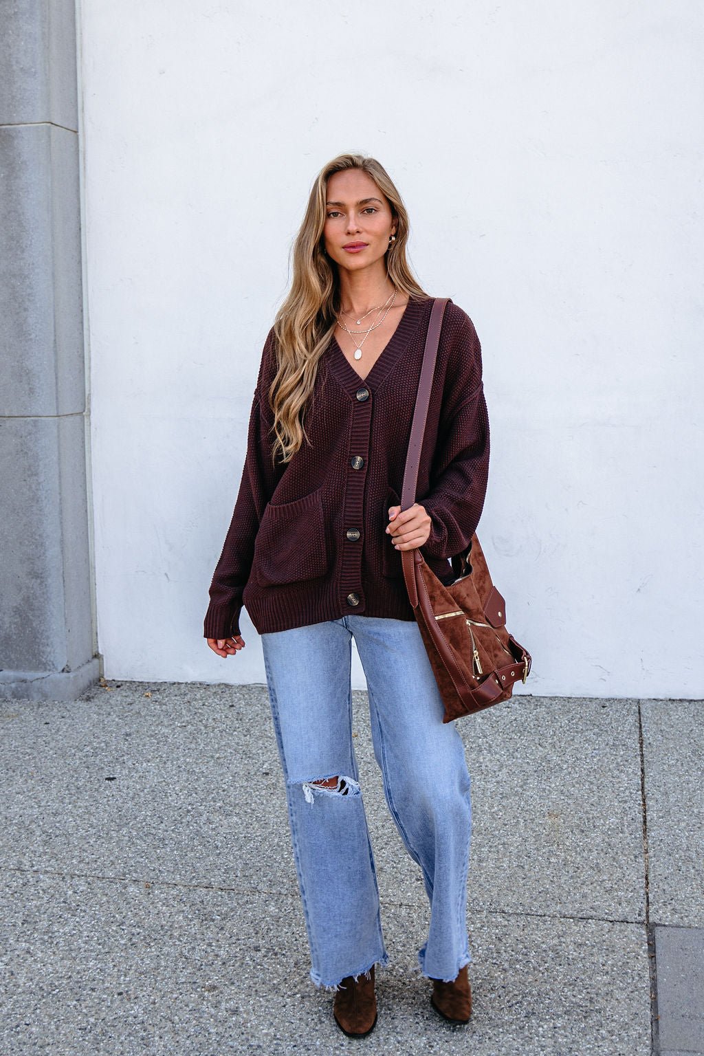 Woman with long hair in an oversized Dark Brown Button Down Cardigan, ripped jeans, brown bag, and boots stands against a white wall.