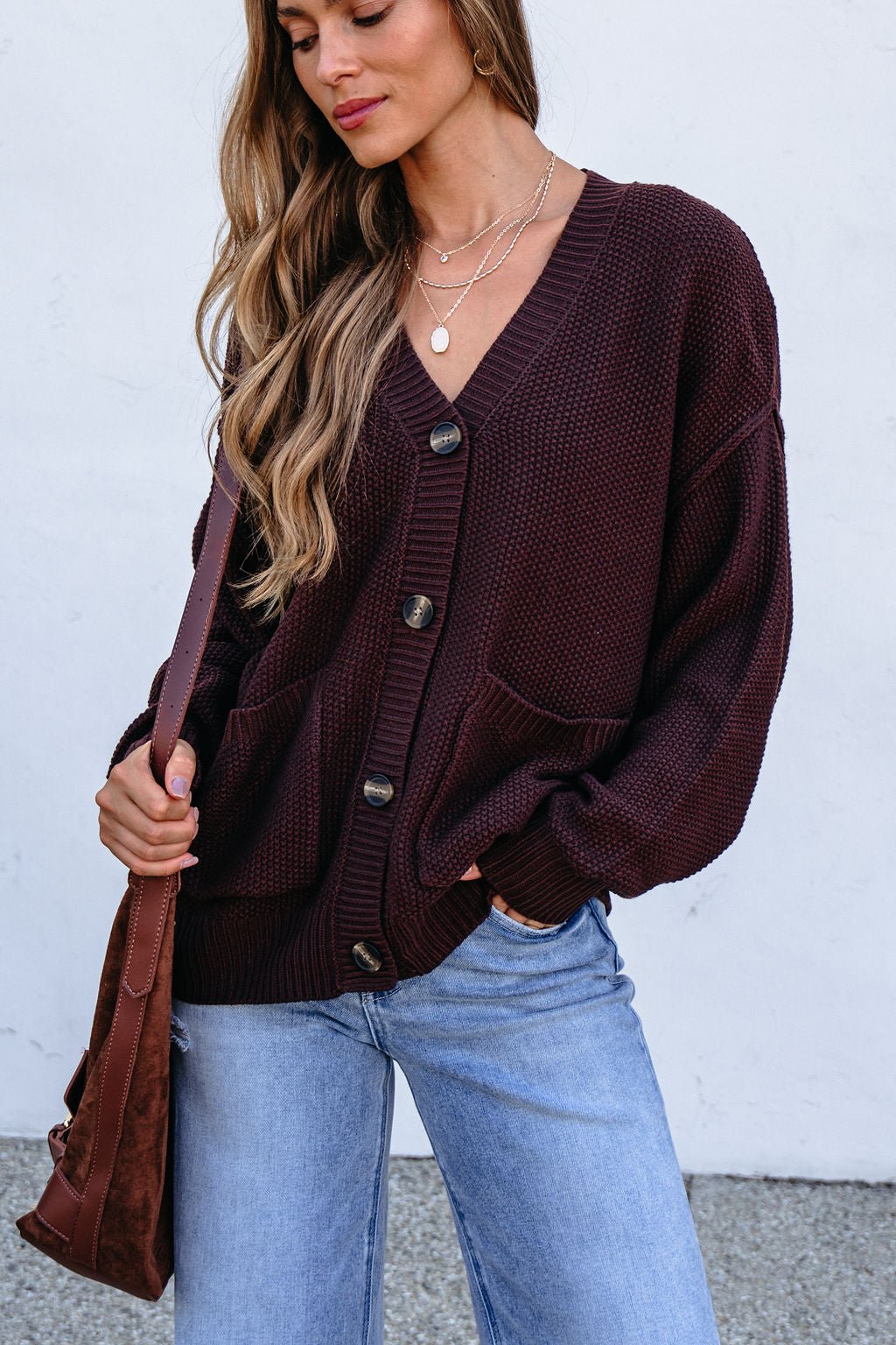Woman wears a Dark Brown Button Down Cardigan, light blue jeans, layered necklaces, and holds a brown bag against a plain background.