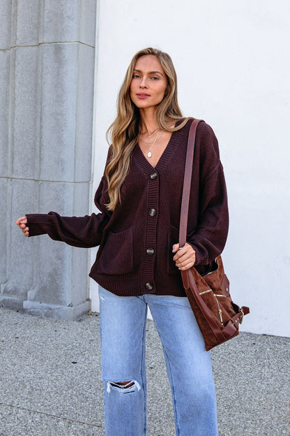 Woman with long hair in a Dark Brown Button Down Cardigan and jeans, holding a brown bag by a stone wall.