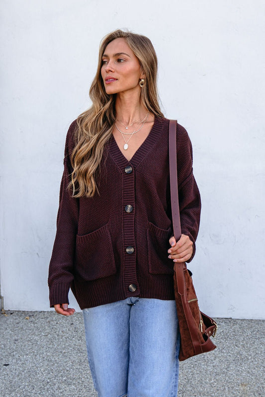 Woman with long hair wears a Dark Brown Button Down Cardigan, light jeans, layered necklaces, and carries a brown bag by a white wall.