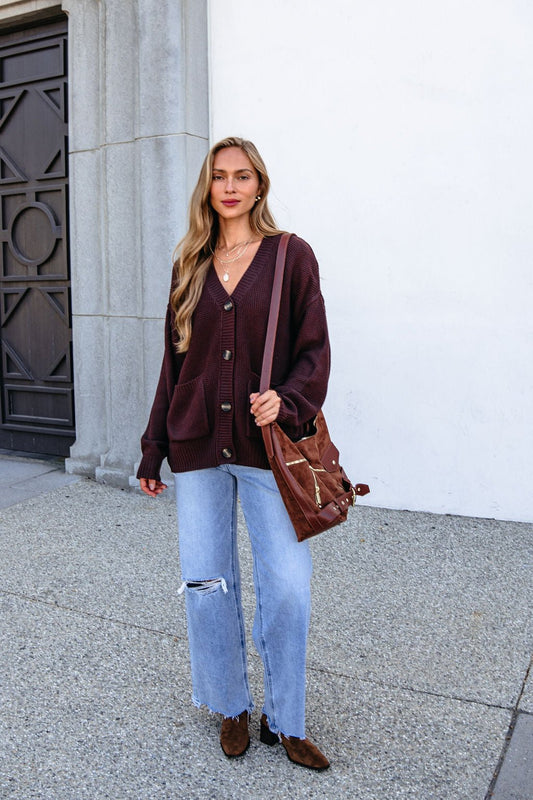 Woman in a Dark Brown Button Down Cardigan and ripped jeans stands outside with a tote bag, wearing brown shoes.