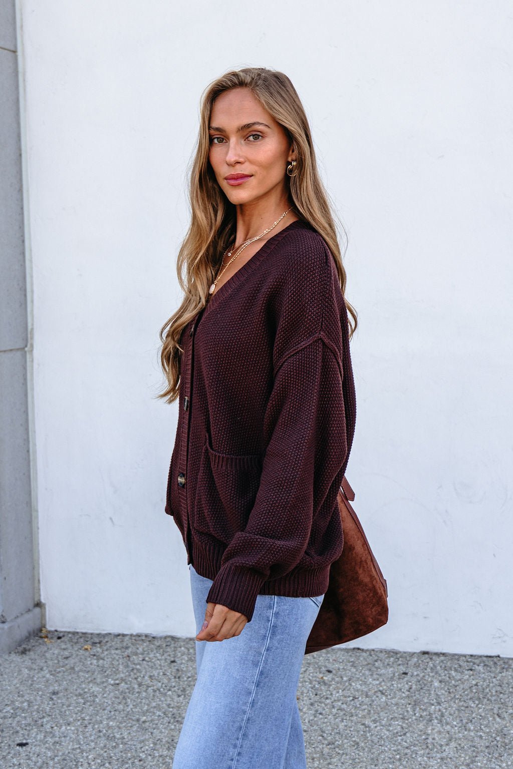 Woman wearing a Dark Brown Button Down Cardigan with light jeans stands outside by a white wall, looking at the camera.