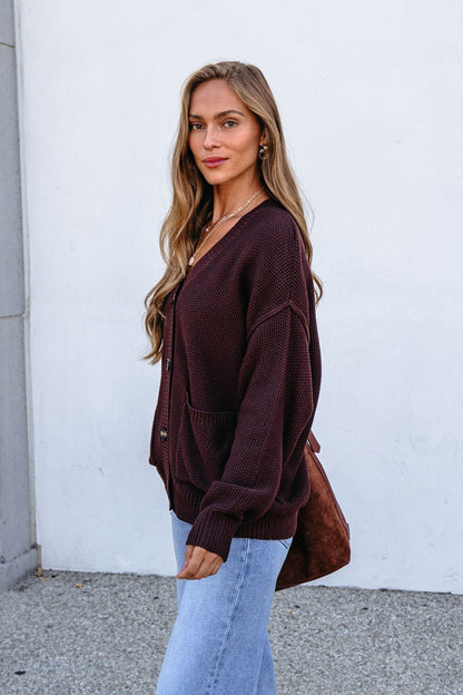 Woman wearing a Dark Brown Button Down Cardigan with light jeans stands outside by a white wall, looking at the camera.