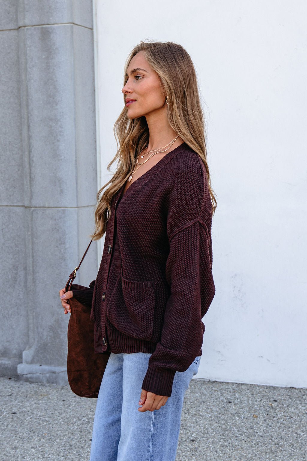 Woman with long hair in a Dark Brown Button Down Cardigan and jeans stands sideways by a stone wall, holding a brown bag.