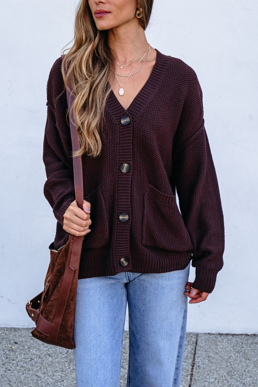 Woman in a Dark Brown Button Down Cardigan, blue jeans, and brown bag stands by a light wall with her long hair loose.