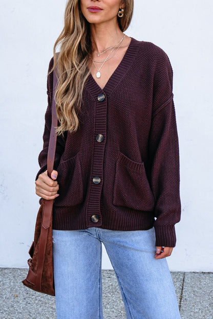 Woman wears a Dark Brown Button Down Cardigan with light blue jeans and a brown bag, standing against a white wall.