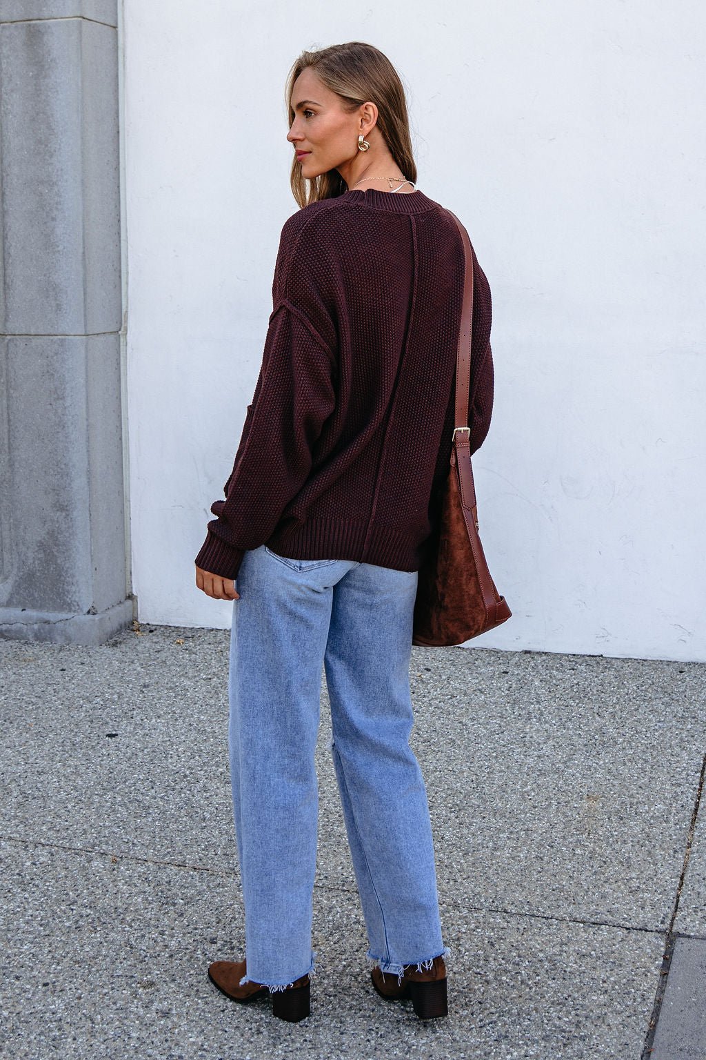 Woman in a dark brown button down cardigan, blue jeans, brown boots, and bag stands outside on a stone walkway, looking over her shoulder.