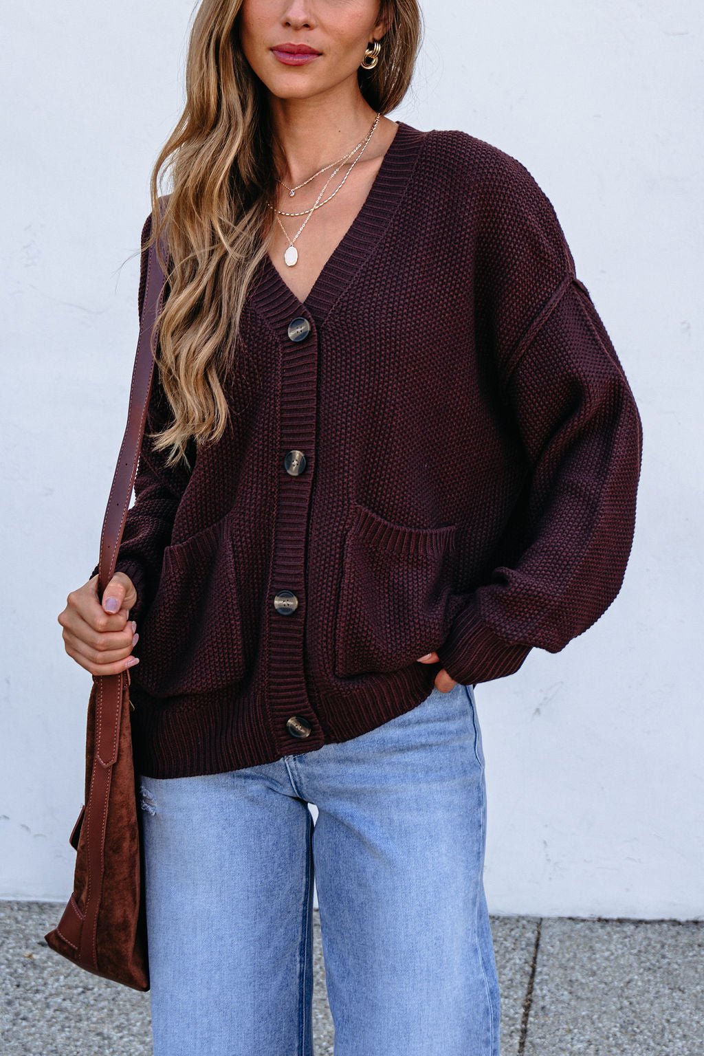 Woman in a dark brown button down cardigan, blue jeans, layered necklaces, and a brown shoulder bag.