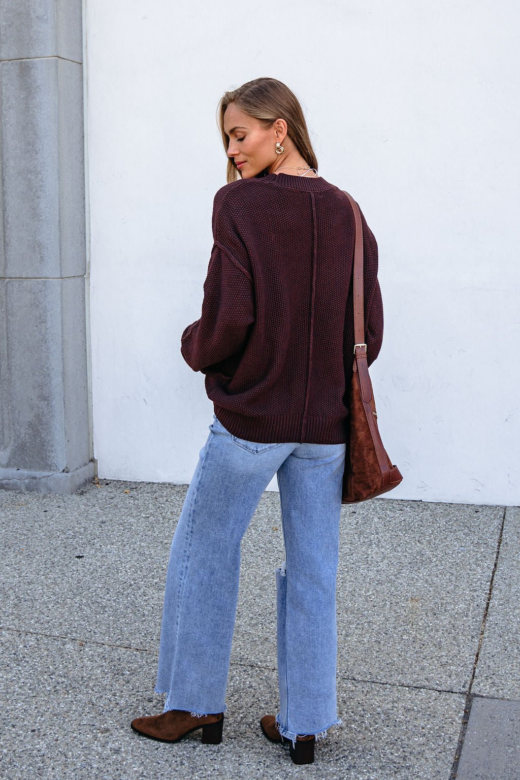 Woman in a dark brown button down cardigan, light blue jeans, and boots stands outside, carrying a matching brown bag over her shoulder.