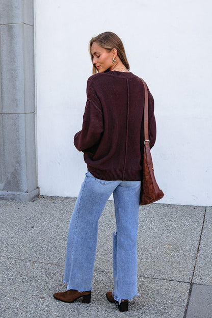 Woman in a dark brown button down cardigan, light blue jeans, and boots stands outside, carrying a matching brown bag over her shoulder.