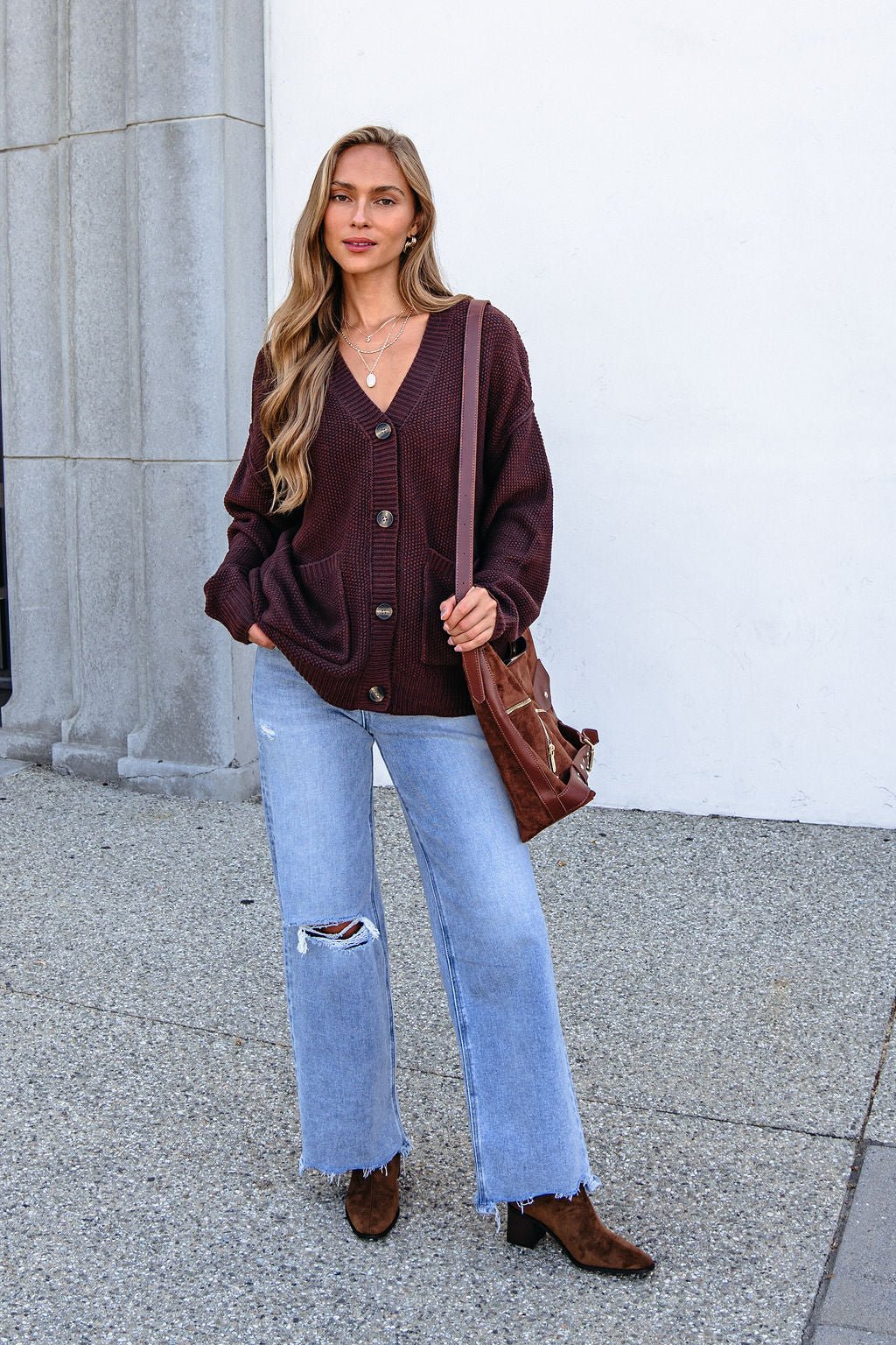 Woman in a Dark Brown Button Down Cardigan, ripped jeans, boots, and bag stands outside near a building.