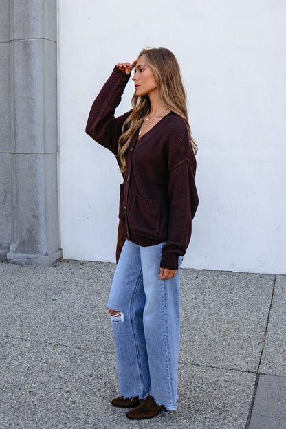 Woman wears an oversized fit dark brown button down cardigan, standing sideways in wide-leg jeans by a white wall.