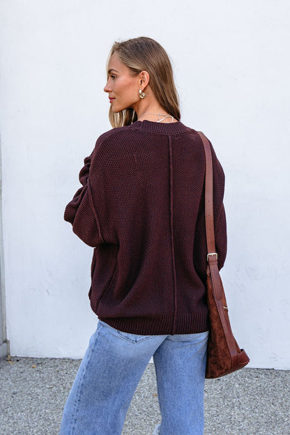Woman with long hair in a dark brown button down cardigan, light blue jeans, and shoulder bag, standing outdoors facing away.