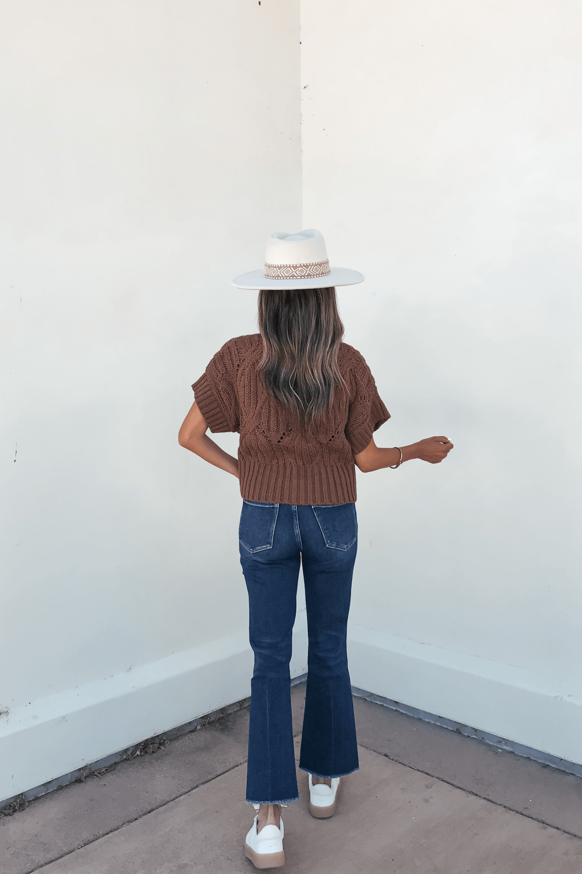Woman stands against a white wall in an autumn staple: a Dark Brown Crochet Knit Sweater, blue jeans, and a white hat.