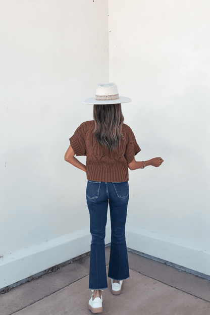 Woman stands against a white wall in an autumn staple: a Dark Brown Crochet Knit Sweater, blue jeans, and a white hat.