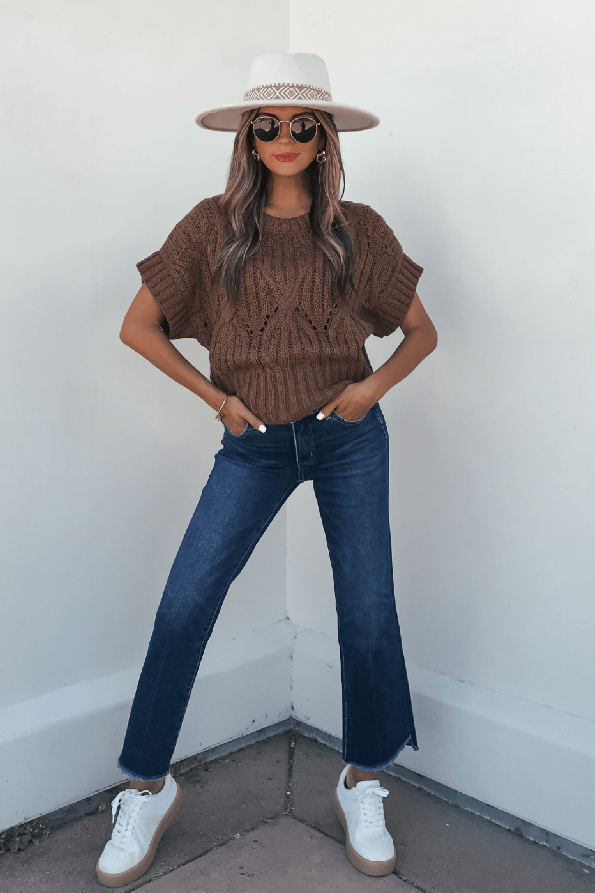 Woman wearing a Dark Brown Crochet Knit Sweater, blue jeans, and accessories stands confidently against a white wall.