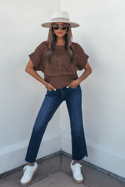 Woman wearing a Dark Brown Crochet Knit Sweater, blue jeans, and accessories stands confidently against a white wall.